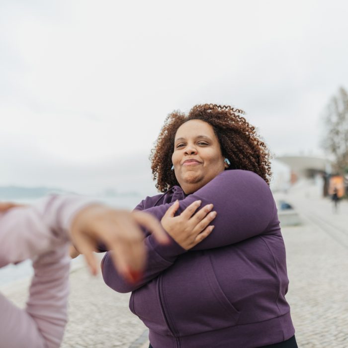 A woman stretches her arms while enjoying a workout session by the beach, showcasing determination and focus amidst a serene landscape of water and clouds.