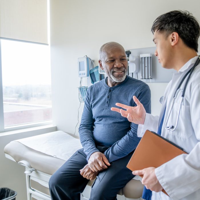 A senior gentleman of African decent sits up on an exam table during a routine medical exam. His doctor is standing to the side of him in a white lab coat as they discuss his health.