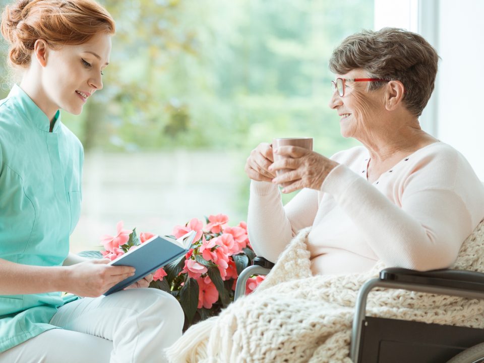 Assistant in blue uniform reads book for elder woman on wheelchair in hospital room