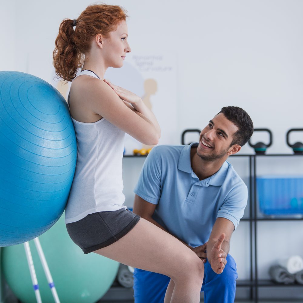 Smiling professional personal trainer helping sportswoman exercising with ball